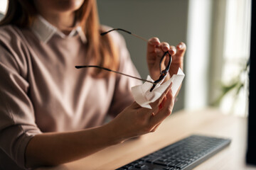 Woman Cleaning Glasses Near Computer in Soft Natural Lighting