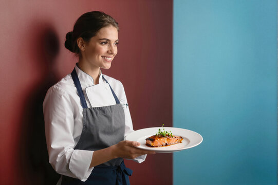 A smiling female chef in a white coat and gray apron holding a plate with salmon dish