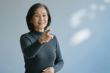 An older Asian woman in a dark sweater pointing forward with her right hand while smiling at the camera against a light blue background.