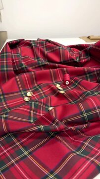 Close-up of gold buttons and a red thread spool falling onto red tartan fabric. Vertical Christmas-themed craft video, ideal for holiday fashion, handmade projects and festive design.