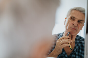 An elderly man studies his reflection in a bathroom mirror, focusing as he sharpens his razor. A...