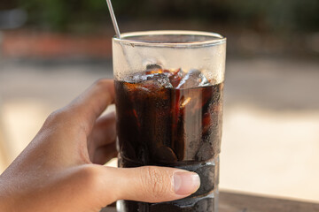 Hand holding a glass of iced black coffee with clear condensation. Symbolizes coolness, refreshment, and summer/hot weather beverage.
