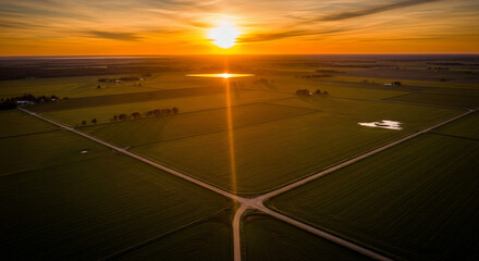 Golden hour aerial view of vast agricultural fields intersected by country roads, bathed in the warm glow of a brilliant sunset on the horizon, creating a serene and expansive rural landscape