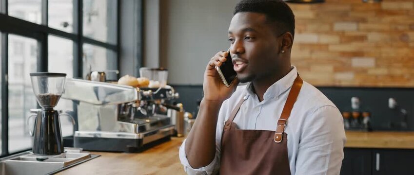 Cafe owner on call: A focused cafe owner, clad in an apron, engages in a phone conversation at his establishment, surrounded by coffee-making equipment. 