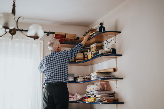 An elderly man stands at a cluttered wall bookshelf, reaching for a book. The room feels warm and lived-in, with papers and decorative items creating a calm, cozy reading atmosphere.