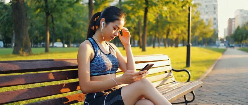 Morning Routine: An athletic woman, engrossed in her phone with earbuds, takes a breather on a park bench bathed in morning sunlight, gearing up for her workout.