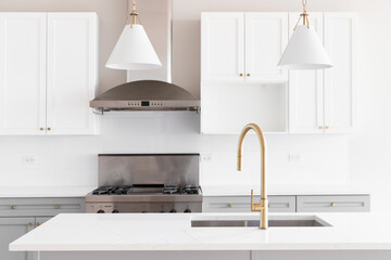 A kitchen detail with white and grey cabinets, gold faucet on a white marble countertop, pendant lights hanging above the island and stainless steel appliances.
