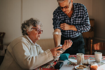 A senior woman sits at a dining table with cookies, tea, and cups while a man in a checkered shirt assists her arm, portraying care, family, and everyday life at home.