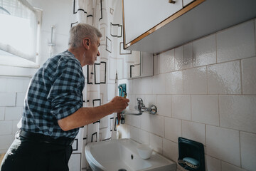 An elderly man in a blue plaid shirt repairs a bathroom faucet at the sink. He holds a tool near the wall taps in a sunlit, tiled bathroom with a patterned shower curtain.