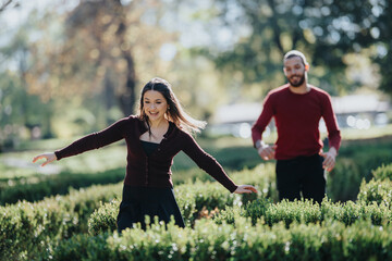 A smiling woman in a dark cardigan dances among trimmed hedges in a sunny park, while a man stands nearby, creating a scene of outdoor activity and carefree, joyful energy.