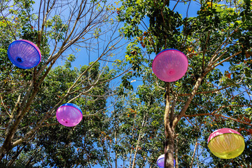 Vibrant Upcycled Lanterns Canopy. Colorful Plastic Food Covers Transformed into Festive Hanging Lamps Amidst Lush Green Tree and Umbrellas. View of Creative Light Fixtures Made From Plastic Food cover