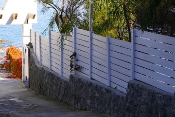 A white fence along a seaside road.
A white wooden fence along a stone wall on a quiet coastal road with a view of the sea and greenery.