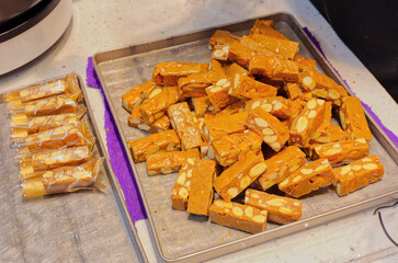 A large batch of freshly made, golden-brown nougat candy packed with nuts is piled on a metal tray, showcasing its chewy, textured appearance, with several pieces individually wrapped to the side.