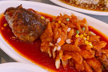A close-up of a large deep-fried fish fillet (or fish head) generously covered in a bright red sweet and sour chili sauce.