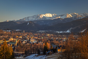 Panoramic view of a town nestled below snow-capped mountains, bathed in the warm glow of the setting sun and clear blue sky. Zakopane town at the foot of the Tatra Mountains in Poland, Europe.
