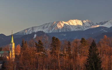 A serene landscape showcases a church steeple, a forest, and majestic, snow-capped mountains basking in the sunlight.  Zakopane town at the foot of the Tatra Mountains in Poland, Europe.
