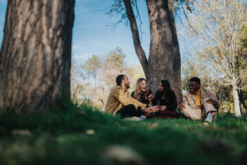 A diverse group of friends sits on the grass beneath tall trees in a sunny park, sharing smiles and conversation. The scene captures friendship, relaxation, and outdoor leisure.