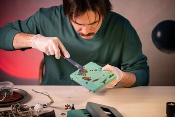 Craftsman renovating electric lamp at worktable, using putty knife tool and scraping off layers of paint from bottom part of metallic base. Upcycling and sustainable living, refurbishing old items.