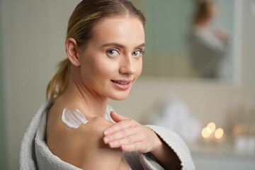 Blond woman with tied-back hair applying shoulder cream in a serene bathroom