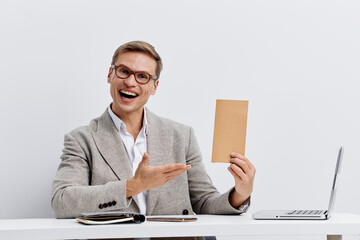 Professional man with glasses in smart casual attire smiling happily while pointing at a blank card in his hand, sitting at a desk with a laptop. Bright, modern studio setting, concept of presentation