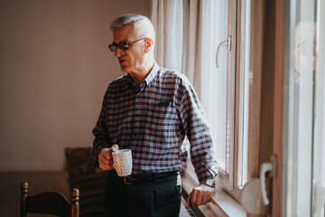 A senior man in a plaid shirt stands by a bright window, savoring a warm coffee. The relaxed indoor moment captures everyday life, quiet reflection, and simple domestic routine.