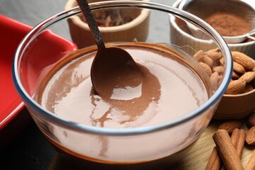 Liquid chocolate dough in bowl, spices, nuts and cocoa powder on table, closeup