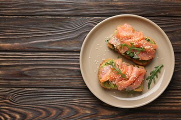 Tasty bruschetta with salmon, avocado, arugula and sesame seeds on wooden table, top view. Space for text