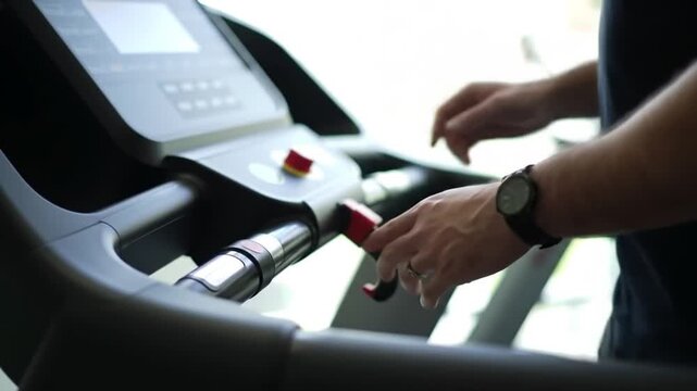 Close-up shot of a person's hands adjusting settings on a treadmill console while wearing a smartwatch, preparing for an indoor cardio workout session at the gym.