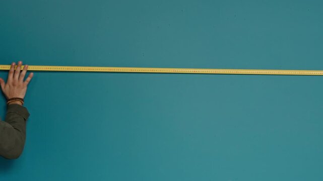 Young man with headband measuring width of teal-coloured wall using yellow flexible ruler while redecorating room