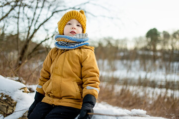 Smiling child in mustard jacket and knit hat crouches in snow, cheeks rosy from the cold. Winter...