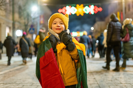 Smiling child in warm clothes wrapped in Lithuanian flag on festive street with lights and crowds at night. Patriotic winter celebration, joyful national holiday atmosphere.