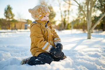 Smiling child in mustard jacket and knit hat crouches in snow, cheeks rosy from the cold. Winter...
