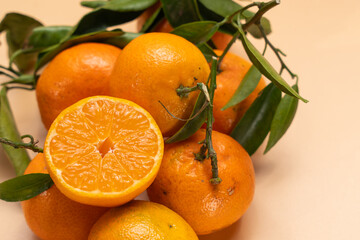 Close-up pile of mandarins with leaves and one sliced half in front, showing juicy pulp. Warm beige background, fresh seasonal citrus concept.