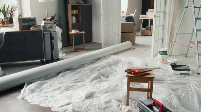 No people view of small wooden stool with handsaw and paper knife placed on it, toolbox, step ladder, paint cans and tray, protective film and large flooring roll in room undergoing renovation