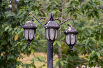 Outdoor post light (street light) with three heads, featuring a black, close-up texture of cast aluminum housing mounted on a tall pole. Provides illumination and security in parks or roads.