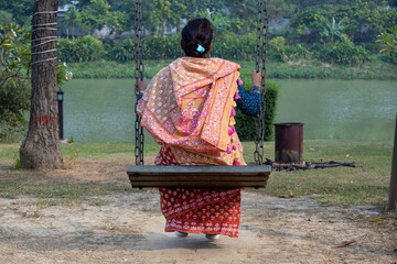 Back view of a Bangladeshi woman in a cotton saree, sitting peacefully on a wooden swing at a resort, overlooking tranquil water and a lush green natural landscape. Relaxation moment.