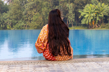 Back view of a Bangladeshi woman in a flowing cotton saree sitting peacefully at the edge of a resort swimming pool, enjoying a quiet moment of relaxation in nature. Travel concept.