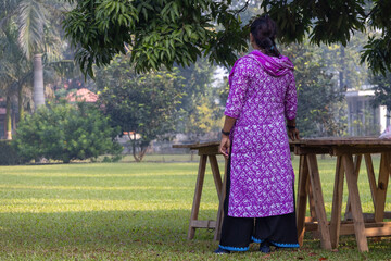 Back view of a solitary Bangladeshi girl wearing a vibrant purple Salwar Kameez, standing alone on a grass field in a park-like setting, looking contemplatively into the distance.