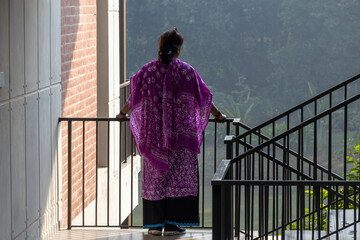 Back view of a Bangladeshi girl standing on a modern apartment balcony, wearing a purple, printed traditional South Asian outfit, commonly known as a Salwar Kameez or three-piece suit.