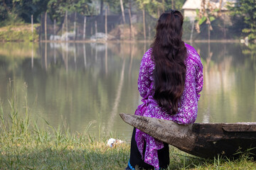 A solitary young Bangladeshi girl in traditional Salwar Kameez clothing sits on the stern of a weathered wooden boat, looking out over a calm river or resort lake in Bangladesh. Back view.