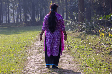 A young Bangladeshi girl in traditional Salwar Kameez outfit walking along a textured brick path, surrounded by vibrant green lawn and dense trees at a tourist resort. Back view.