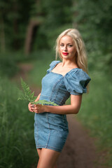 Fototapeta premium Young woman with long blonde hair wearing a denim dress stands on a path in a lush green forest, holding a fern, embodying natural beauty and serenity in nature