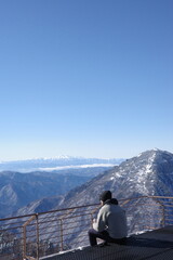 Person sitting on mountain viewpoint overlooking vast alpine landscape under clear sky