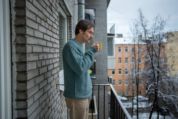 Thoughtful man drinking hot coffee on balcony breathing fresh winter air. Relaxed slow morning, mindful pause, peaceful solitude. Pensive male standing on apartment balcony with warm mug in hands.