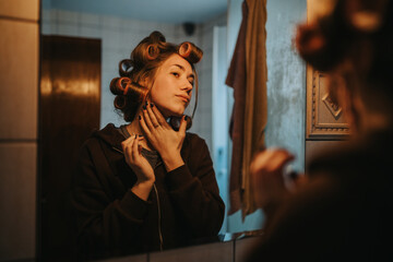 A young woman stands at a bathroom mirror with hair in curlers, wearing a hoodie, as she touches her neck and applies makeup in a casual at-home moment. © qunica.com
