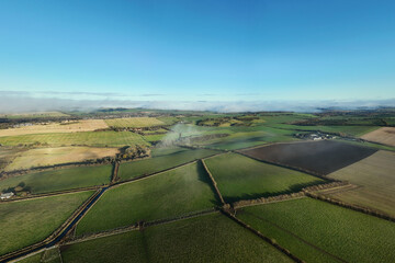 Aerial view of green farmland and rolling hills near Beecraigs Country Park in West Lothian, Scotland, captured on a clear sunny day with soft morning mist over the fields.