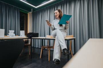 A young woman in comfortable clothing sits on a table in a modern collaboration center, using her phone and holding a clipboard. Relaxed, focused, and ready to brainstorm.