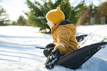Laughing child in mustard jacket and yellow hat rides in a snow shovel, sitting low in the snow. Improvised winter sledding, playful spontaneity in cold sunlight.