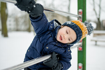Child in winter clothes climbs metal bars on a snowy playground, focused and active. A cold day scene of movement and exploration in a city park.