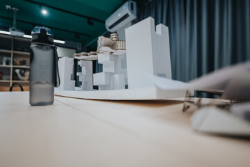 A scale architectural model built from white blocks and cardboard sits on a wooden table in a bright studio. The scene emphasizes creativity, teamwork, and planning in a collaborative design space.